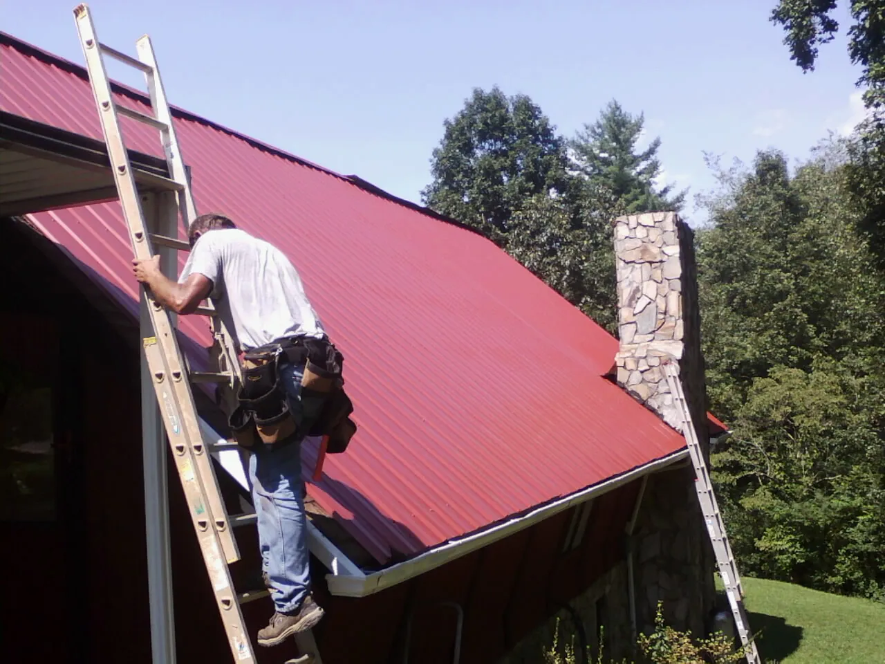 Roofer on a ladder beside a red metal roof and stone chimney — Soffit & Fascia Repair by Roofing Brothers in Caseyville
